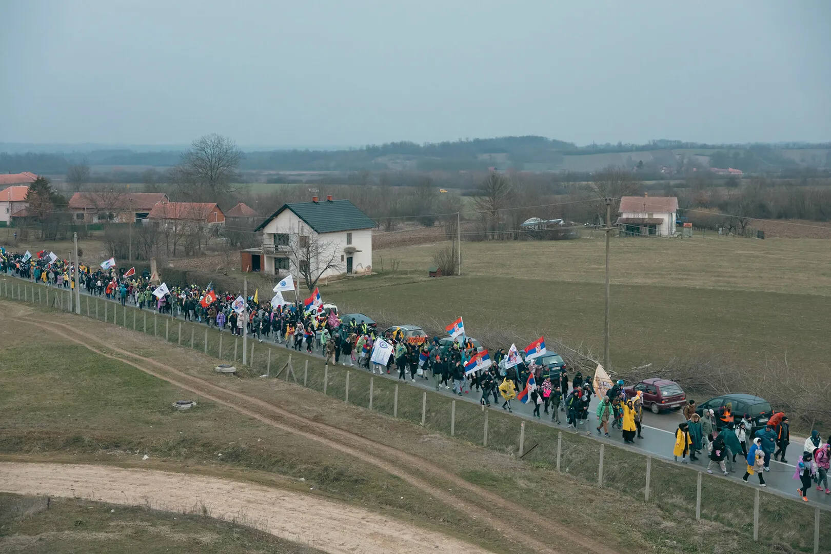 A varied line of students passing through a village. Surrounded by grass and village roads, happily waving flags.