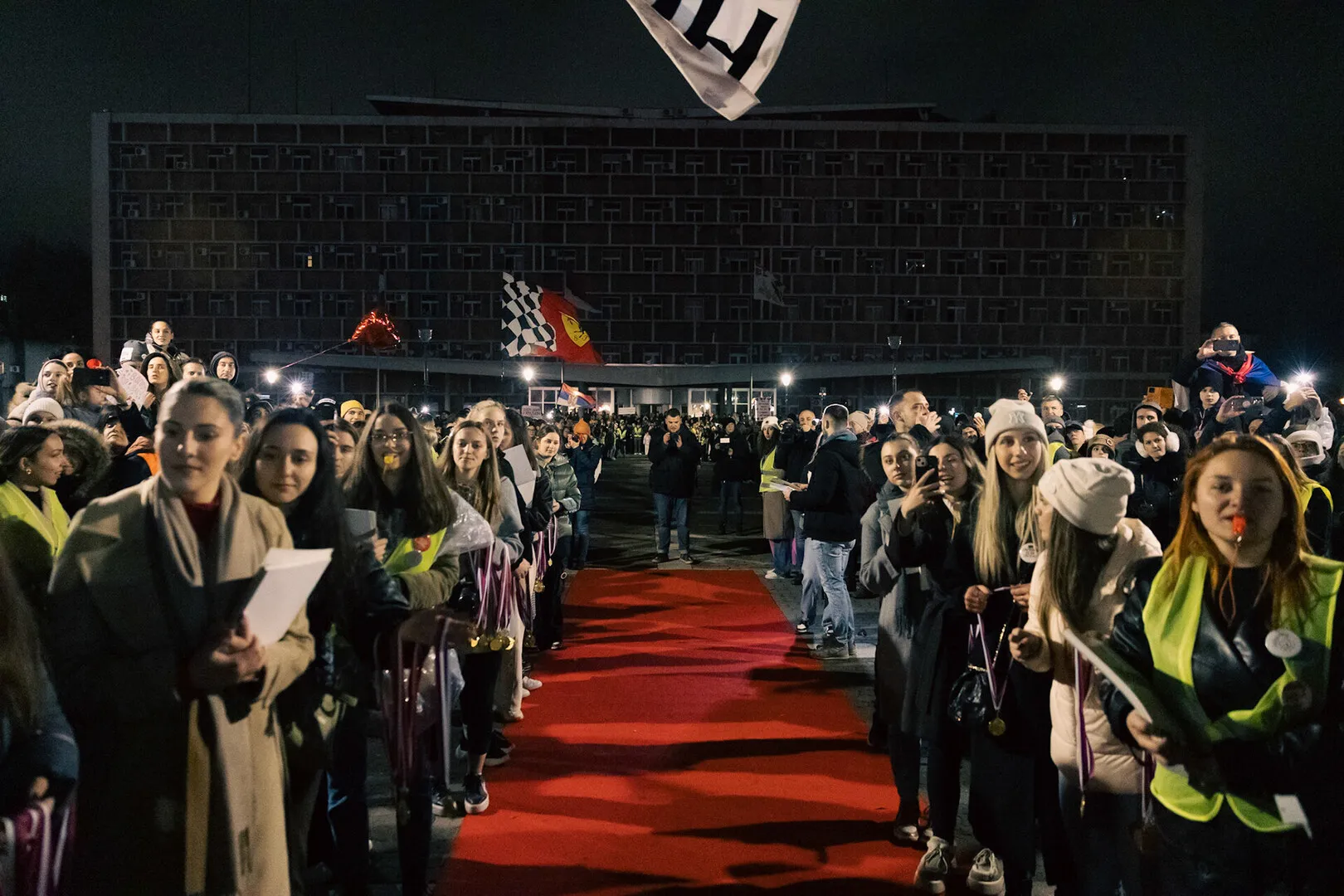 A red carpet, surrounding by a great number of citizens, readily awaits the steps of the walkers after a difficult journey.